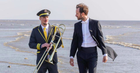 Dvaja muži, jedne v obleku, druhý v armádnej hudobnej uniforme s hudobným nástrojom, sa prechádzajú na pobreží pri mori